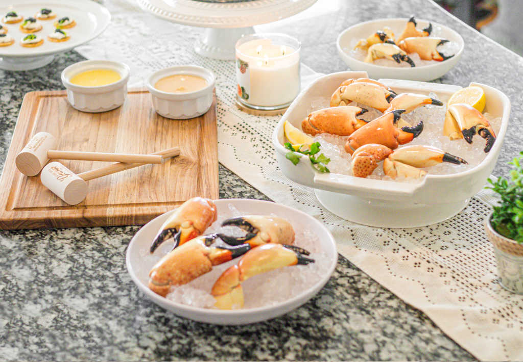 A spread of stone crab claws on ice served in white bowls, accompanied by dipping sauces, wooden crab mallets, and garnished with lemon and herbs on a lace-covered table
