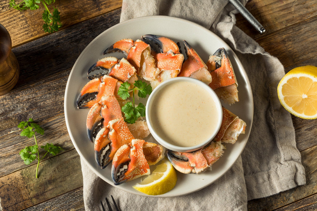 A plate of stone crab claws with mustard dipping sauce, garnished with fresh parsley and lemon wedges. The dish is set on a rustic wooden table with a beige napkin, a fork, and a pepper grinder nearby.