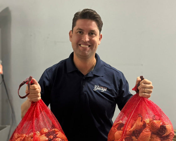 A smiling man in a navy blue polo shirt with a 'Grass' logo holds two large red mesh bags filled with orange-red grass clippings in a warehouse setting.
